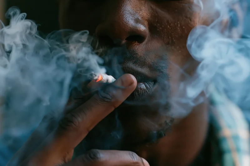 Young black man smoking a cigarette, close-up on lower face and hand, smoke swirling around his mouth