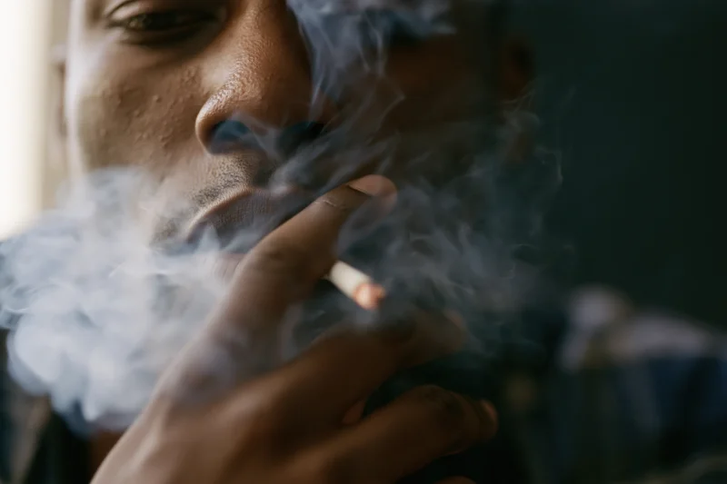 Young black man smoking a cigarette, exhaling smoke, close-up of lower face and hand