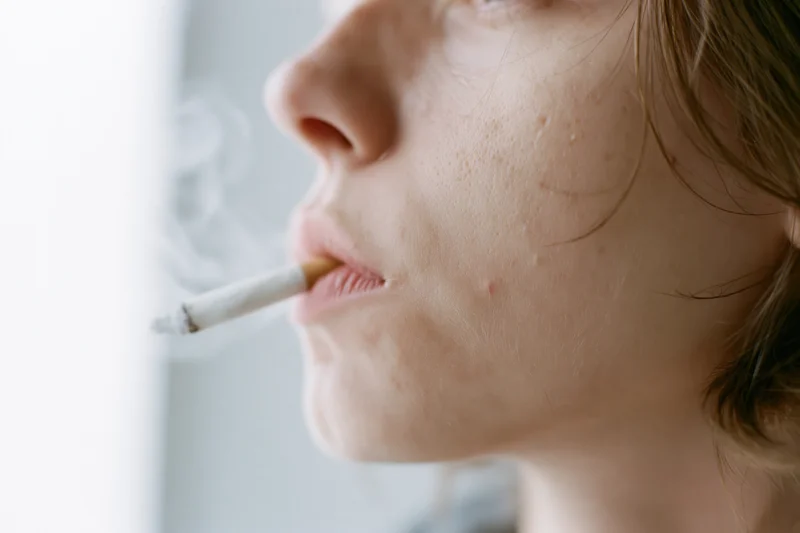 Young woman in profile smoking a cigarette indoors, exhaling smoke near a window