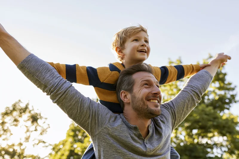 Father carrying young son on his shoulders outdoors with arms raised, both smiling