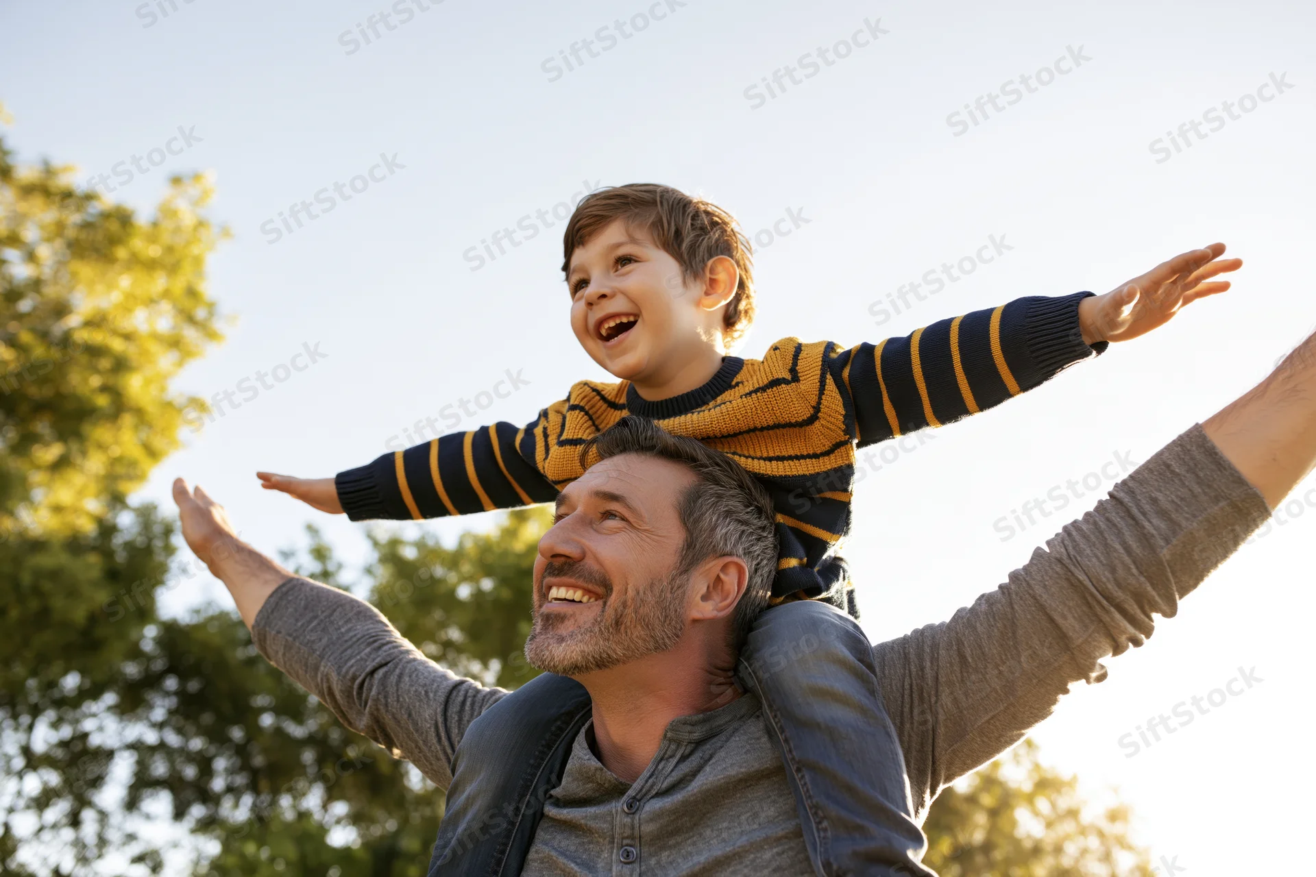 Father carrying young son on his shoulders outdoors with arms outstretched, both smiling