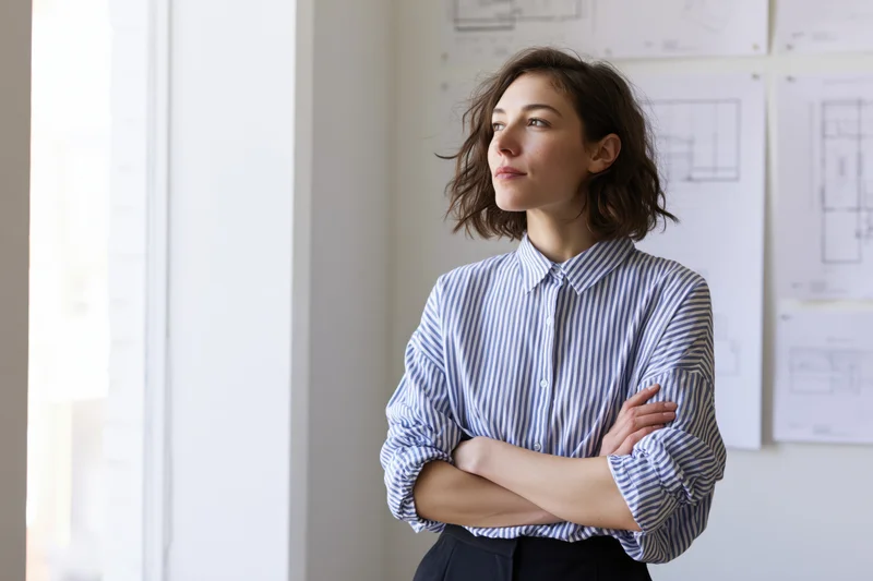 Woman architect standing with arms crossed in her studio looking thoughtfully toward the window