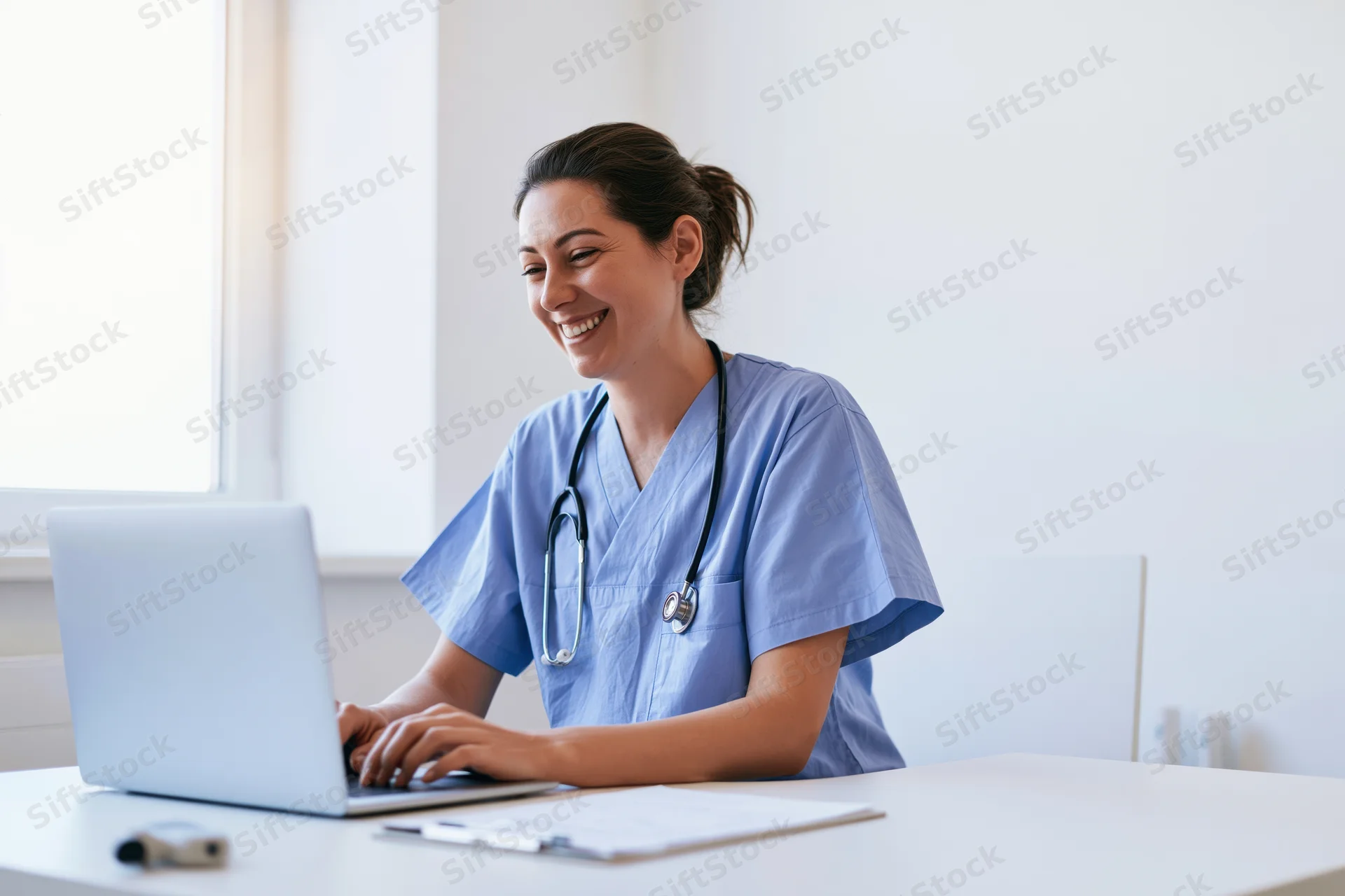 Female doctor in blue scrubs typing on laptop at desk in bright office