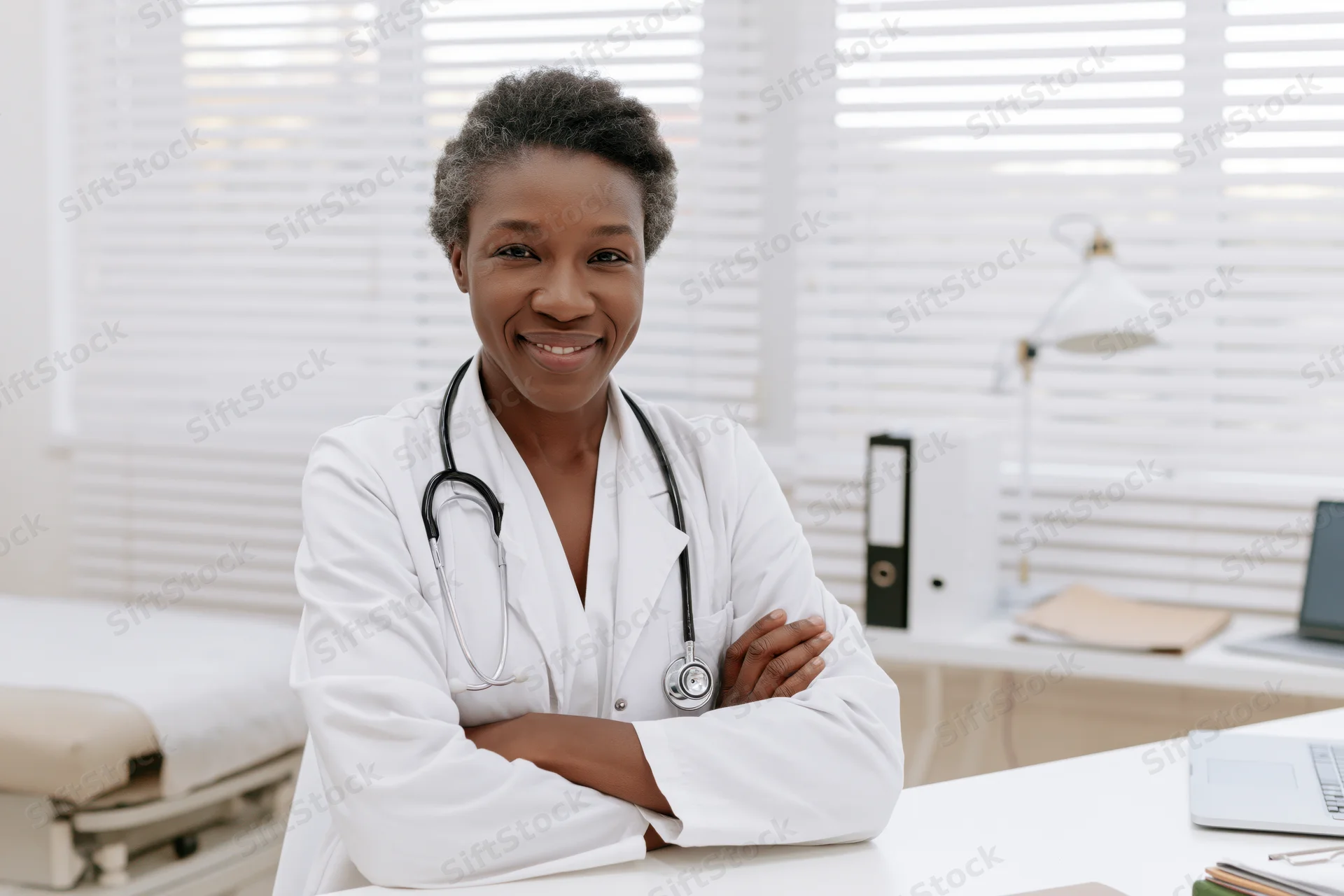 Smiling female doctor in white lab coat with stethoscope around her neck, arms crossed, standing in a bright clinic office