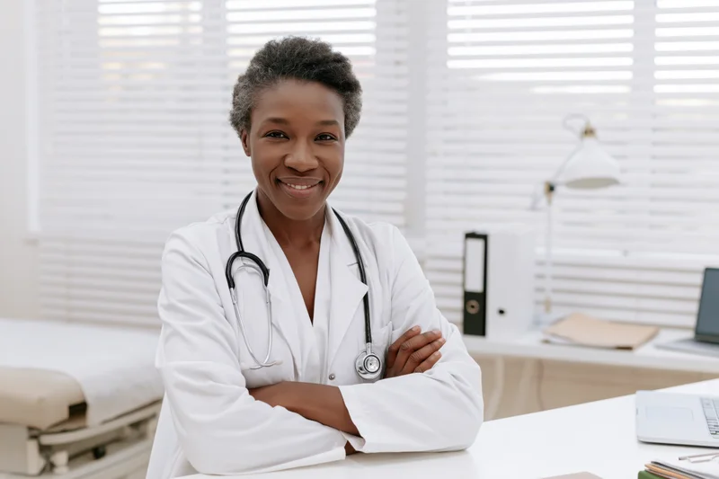 Smiling female doctor in white lab coat with stethoscope around her neck, arms crossed, standing in a bright clinic office