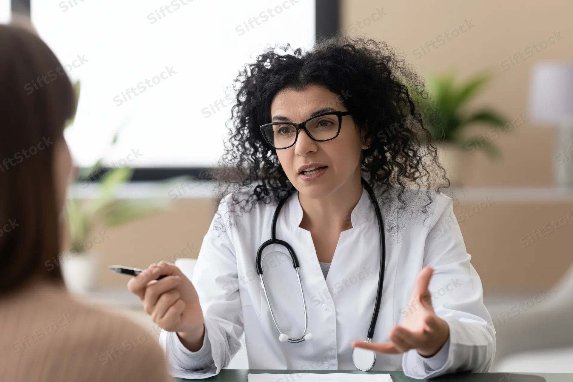 Female doctor in white coat with stethoscope speaking to patient at desk in clinic