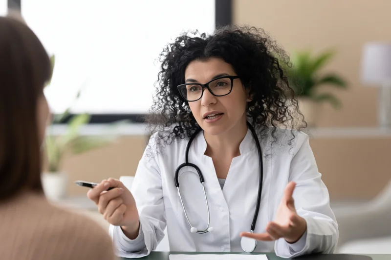 Female doctor in white coat with stethoscope speaking to patient at desk in clinic