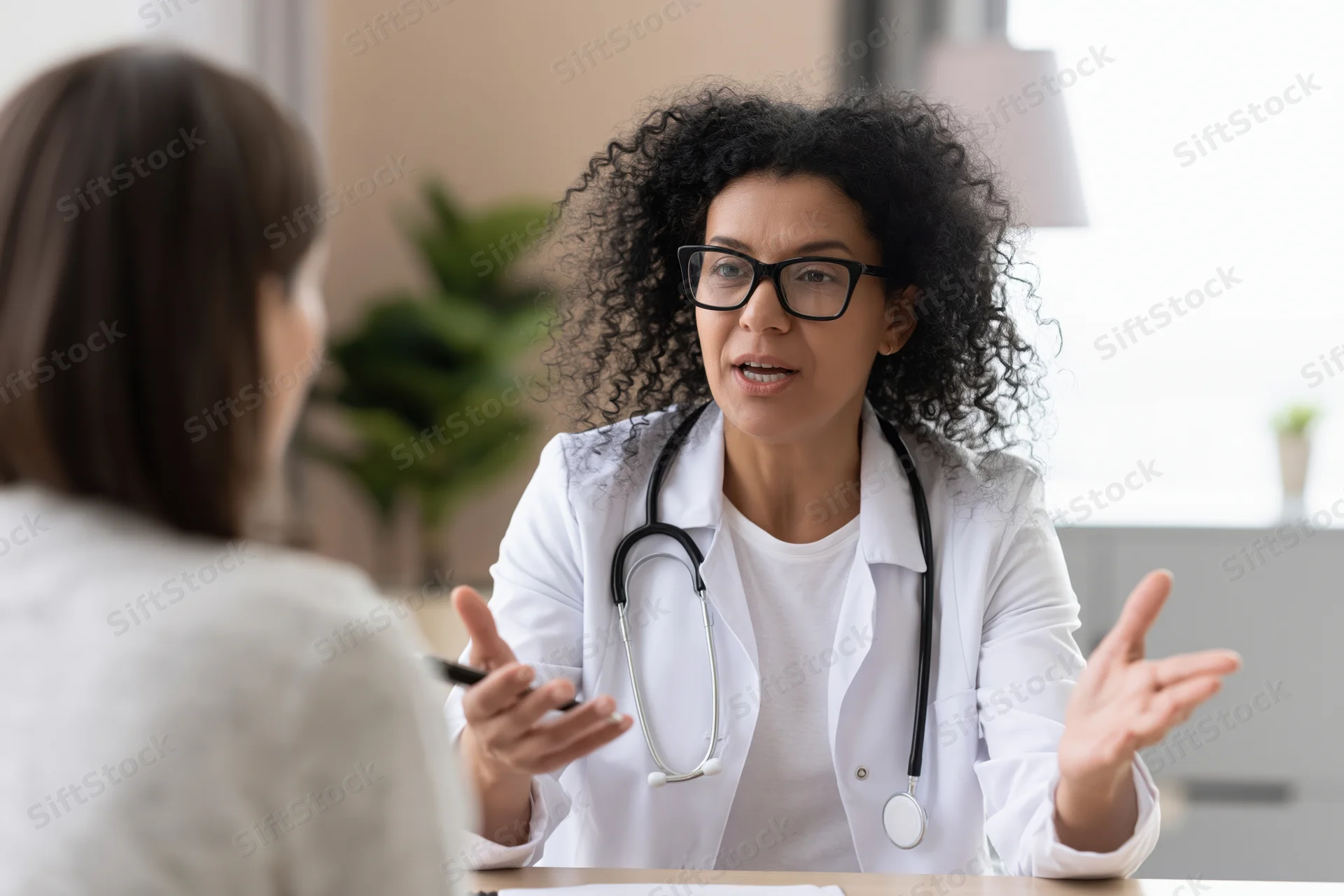 Female doctor in white coat with stethoscope talking and gesturing during consultation with patient in medical office