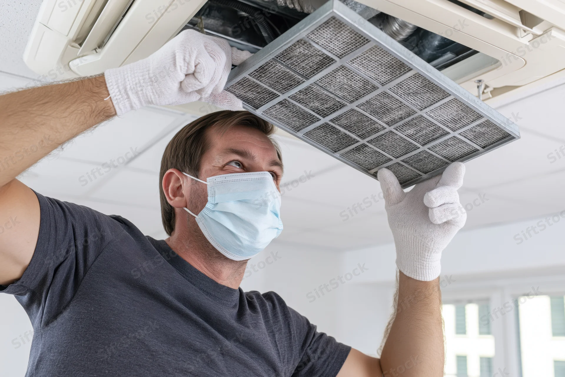 Hvac technician removing air filter from ceiling-mounted unit while wearing protective mask and gloves