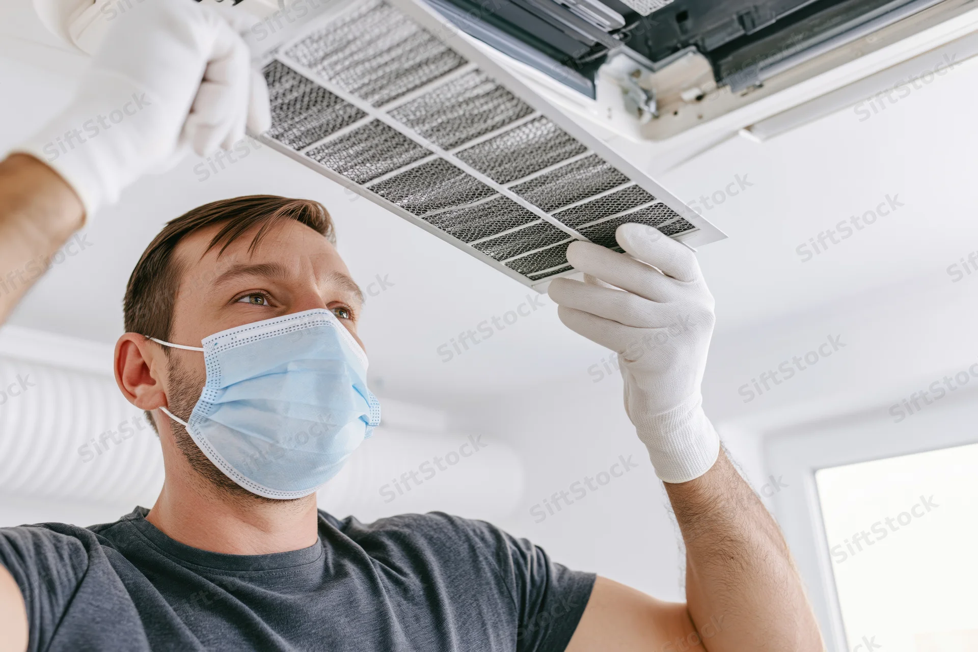 Hvac technician in white gloves and blue mask removing air filter from ceiling unit