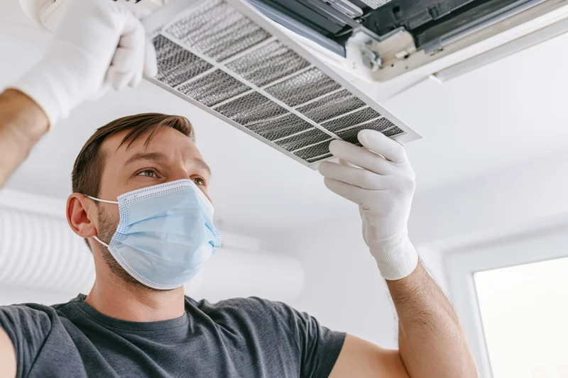 Hvac technician in white gloves and blue mask removing air filter from ceiling unit