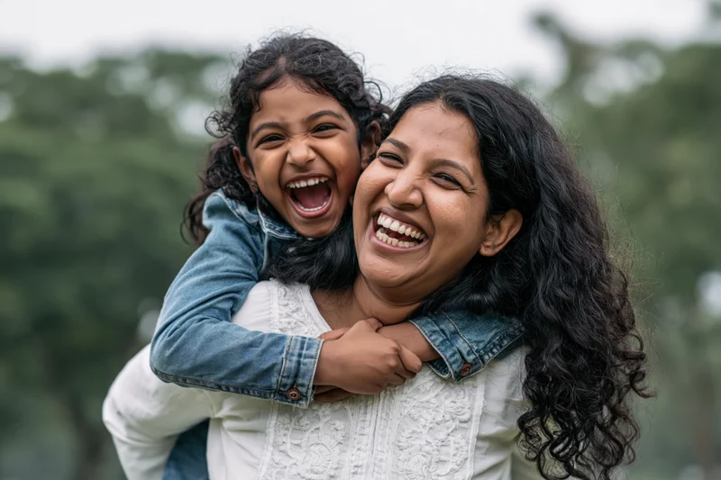 Indian mother giving young daughter a piggyback ride outdoors, both laughing joyfully