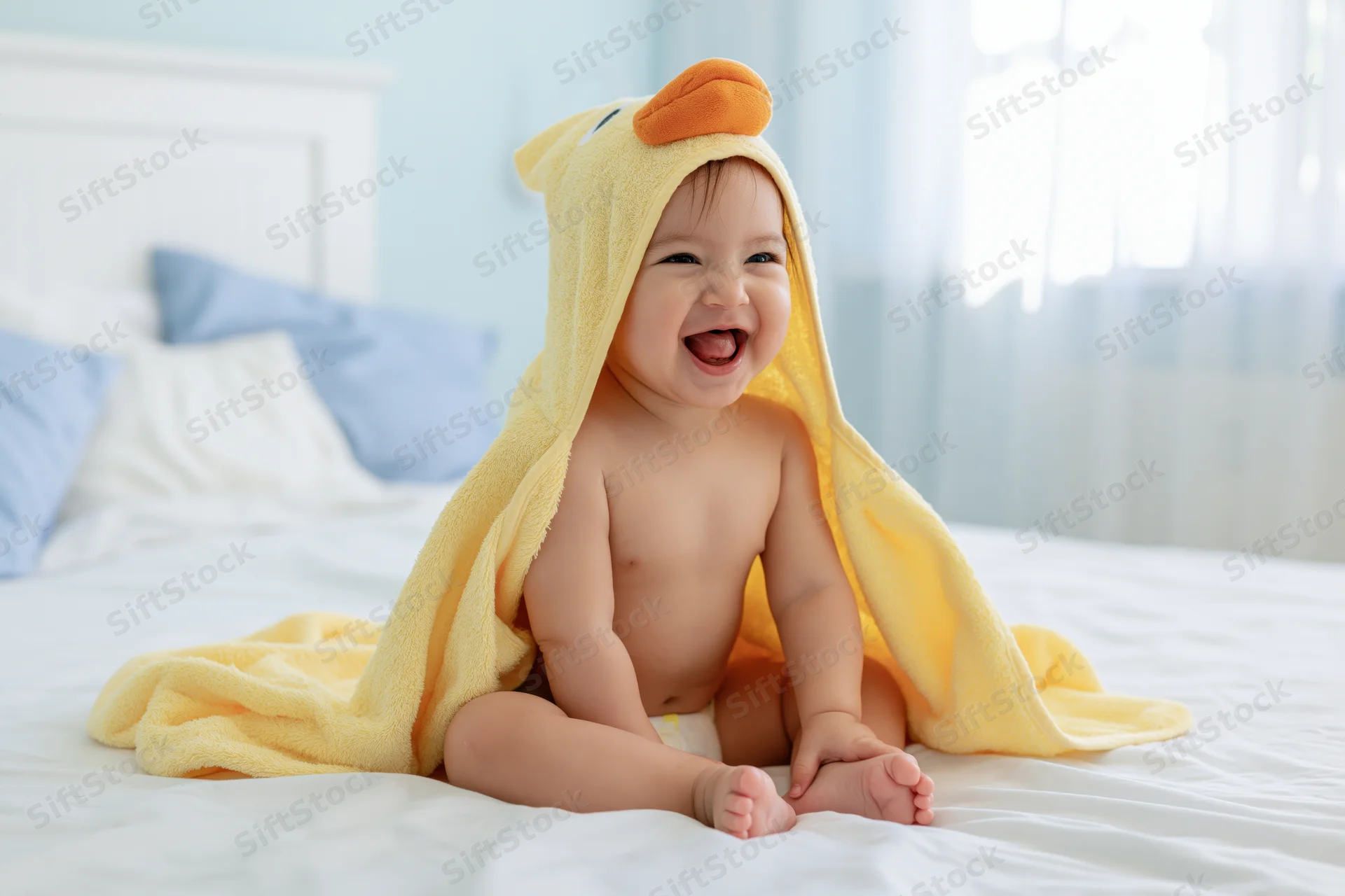 Laughing baby sitting on white bed wearing yellow hooded towel