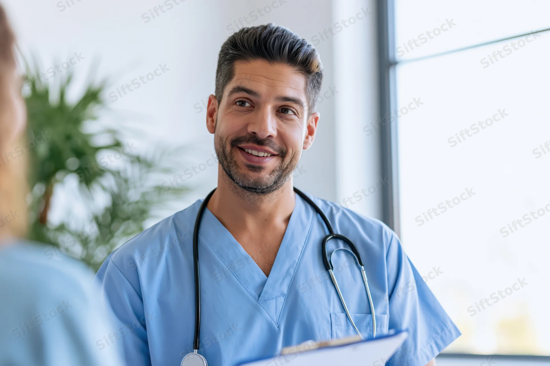Male doctor in blue scrubs smiling while consulting with a patient in a clinic office
