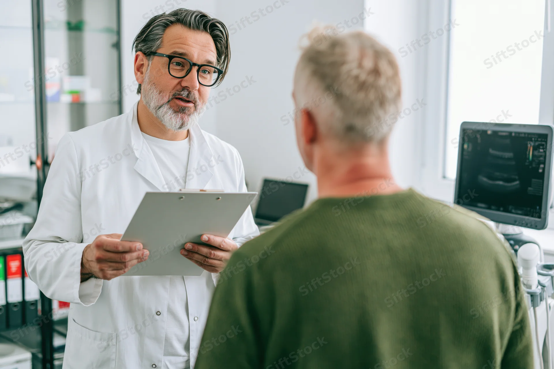 Male doctor in white coat holding clipboard during consultation with patient in clinic