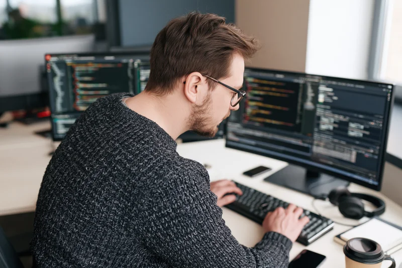 Male programmer typing code on dual monitors at office desk