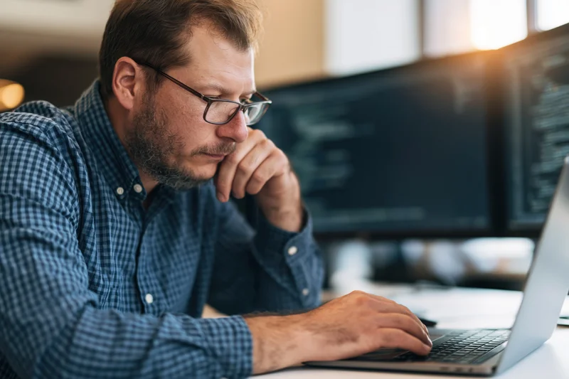 Man in checkered shirt and glasses concentrating on laptop at office desk with monitors in background