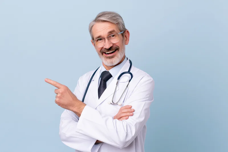 Senior male doctor in white lab coat pointing with smile, arms crossed, against light blue background