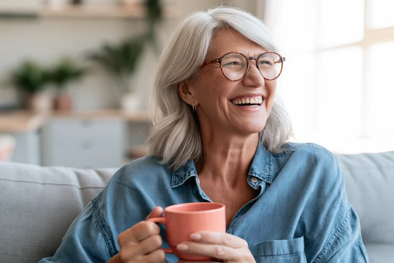 Senior woman laughing while holding a mug on a couch in a bright living room