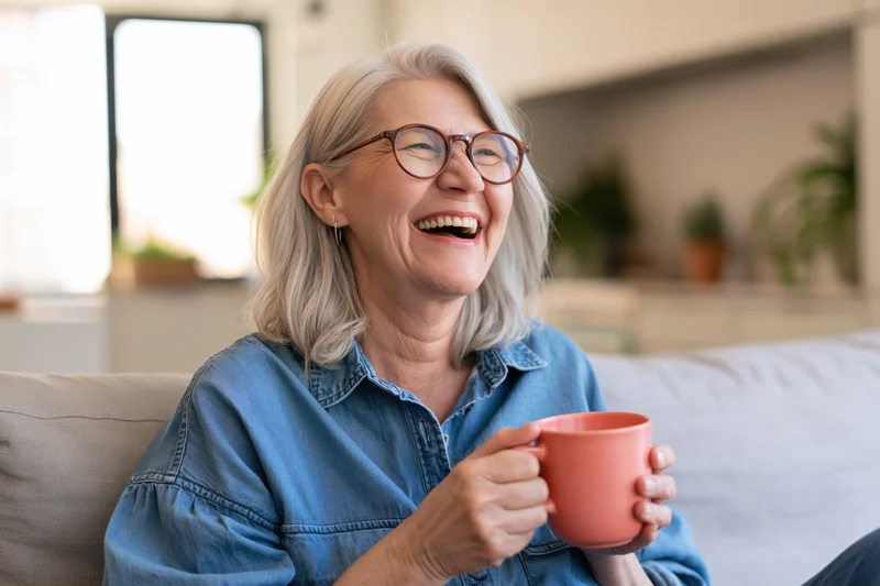 Senior woman laughing while holding a pink mug on a couch in a living room