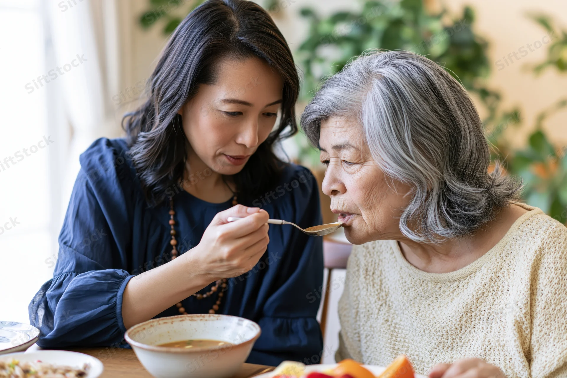 Middle-aged asian woman feeding soup to her senior mother at home dining table