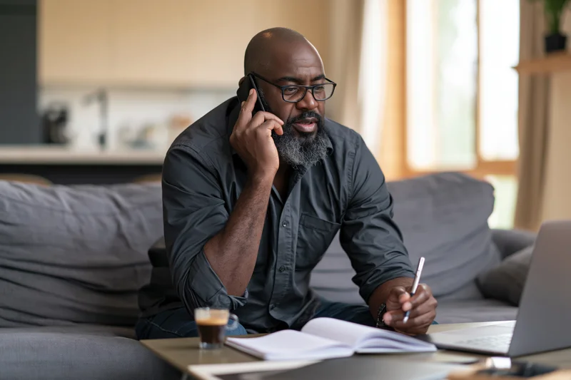 Middle-aged black man talking on phone while working at home on sofa with laptop and notebook