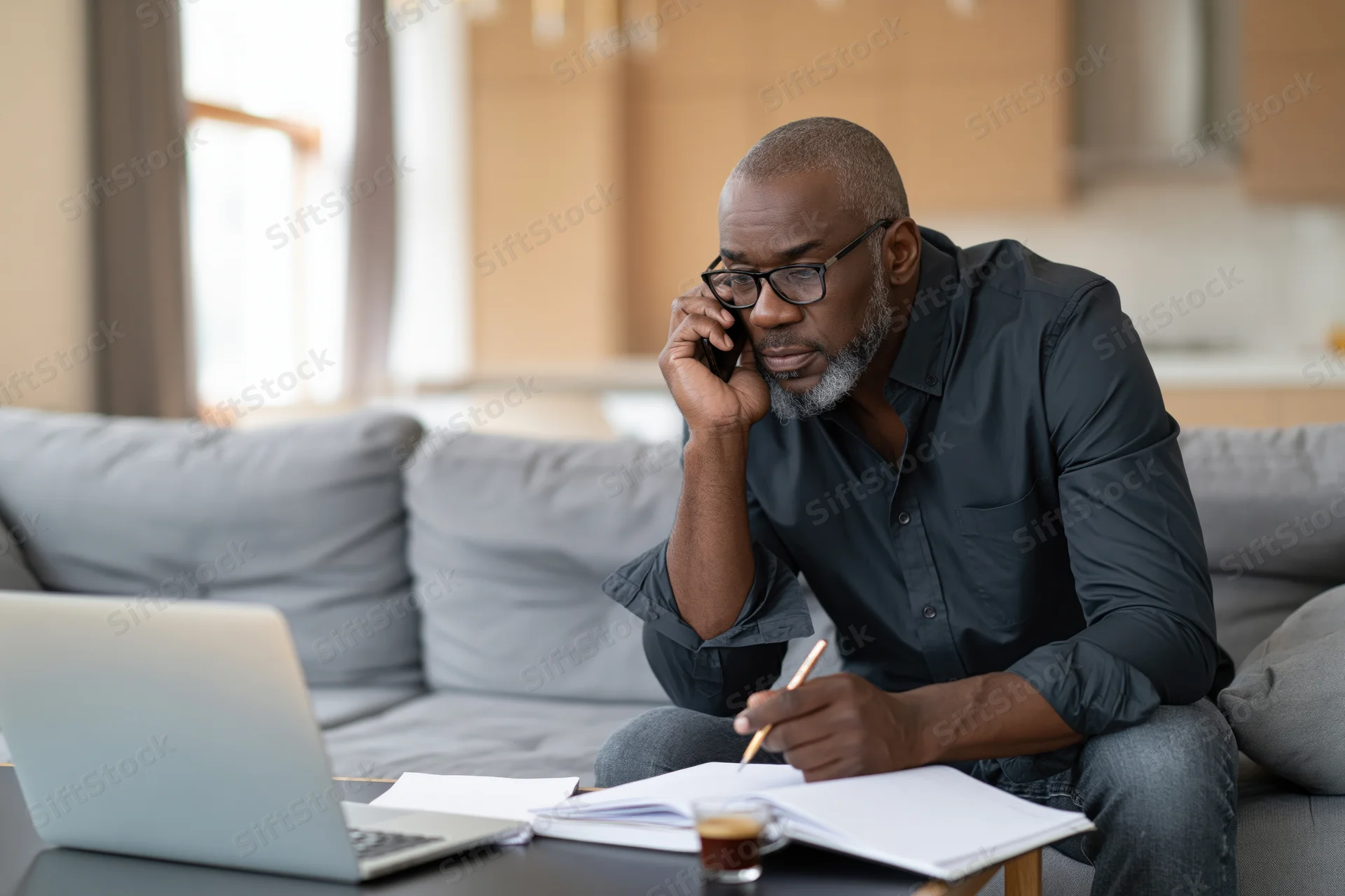 Middle-aged black man talking on phone while writing in notebook at home office setup on couch