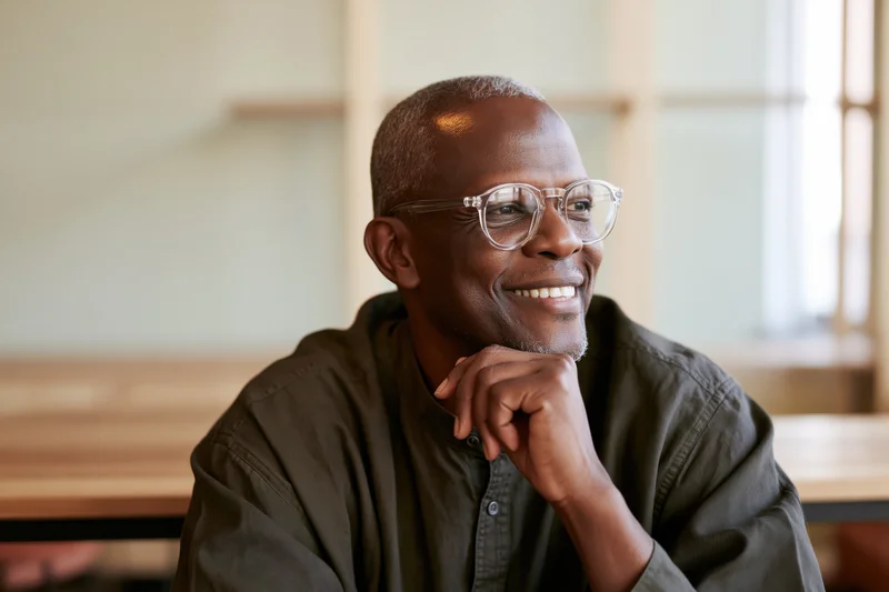 Smiling senior black man with clear-framed glasses resting hand on chin in bright indoor workspace