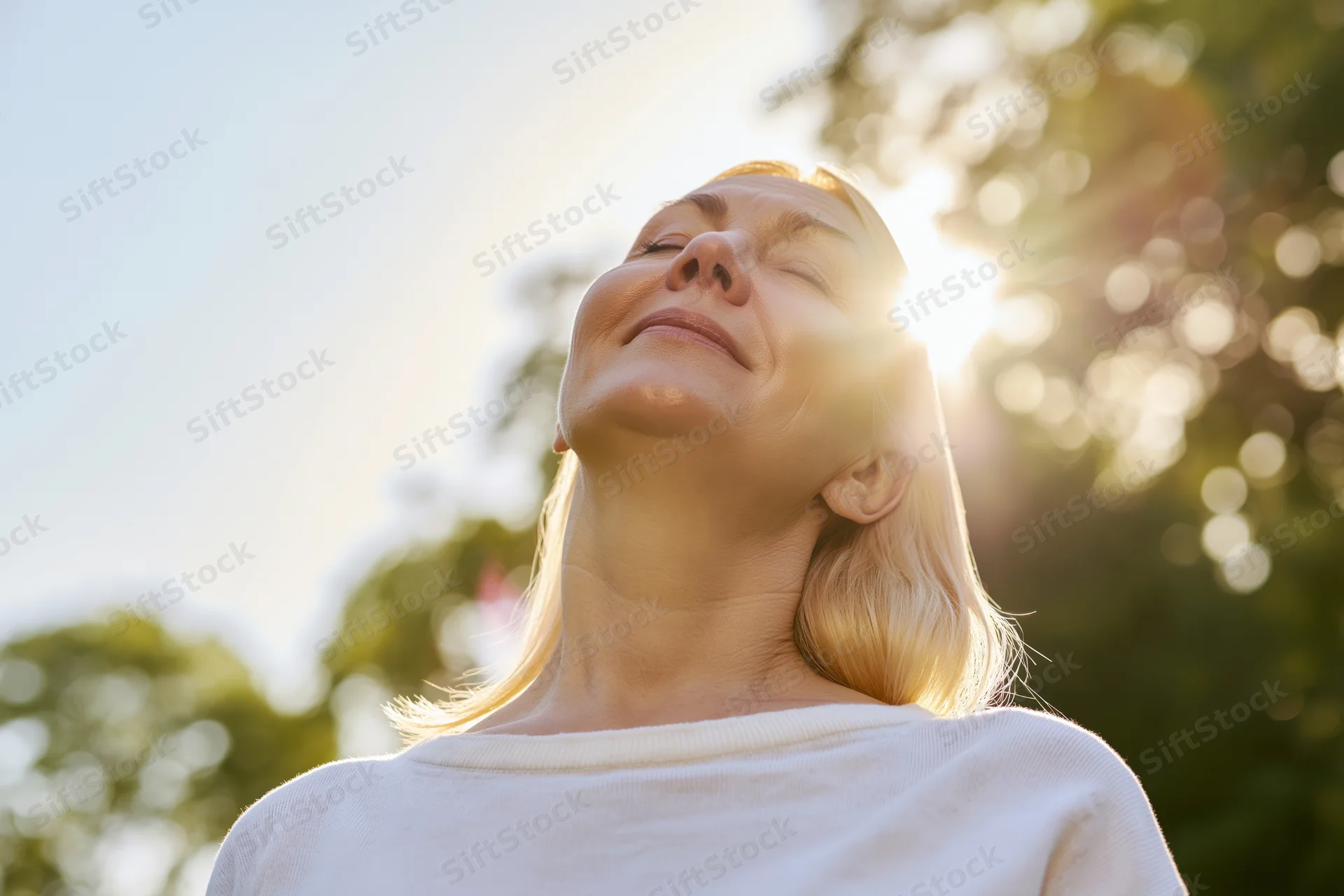 Middle-aged blonde woman tilting head back with eyes closed in sunlight outdoors