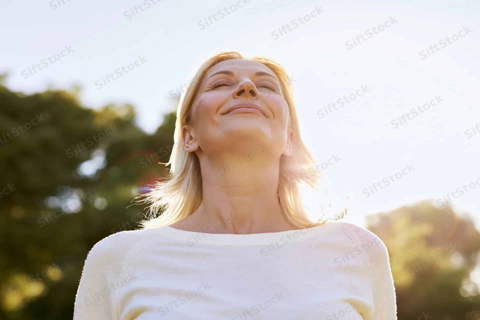 Middle-aged blonde woman tilting head back with eyes closed in sunlight outdoors
