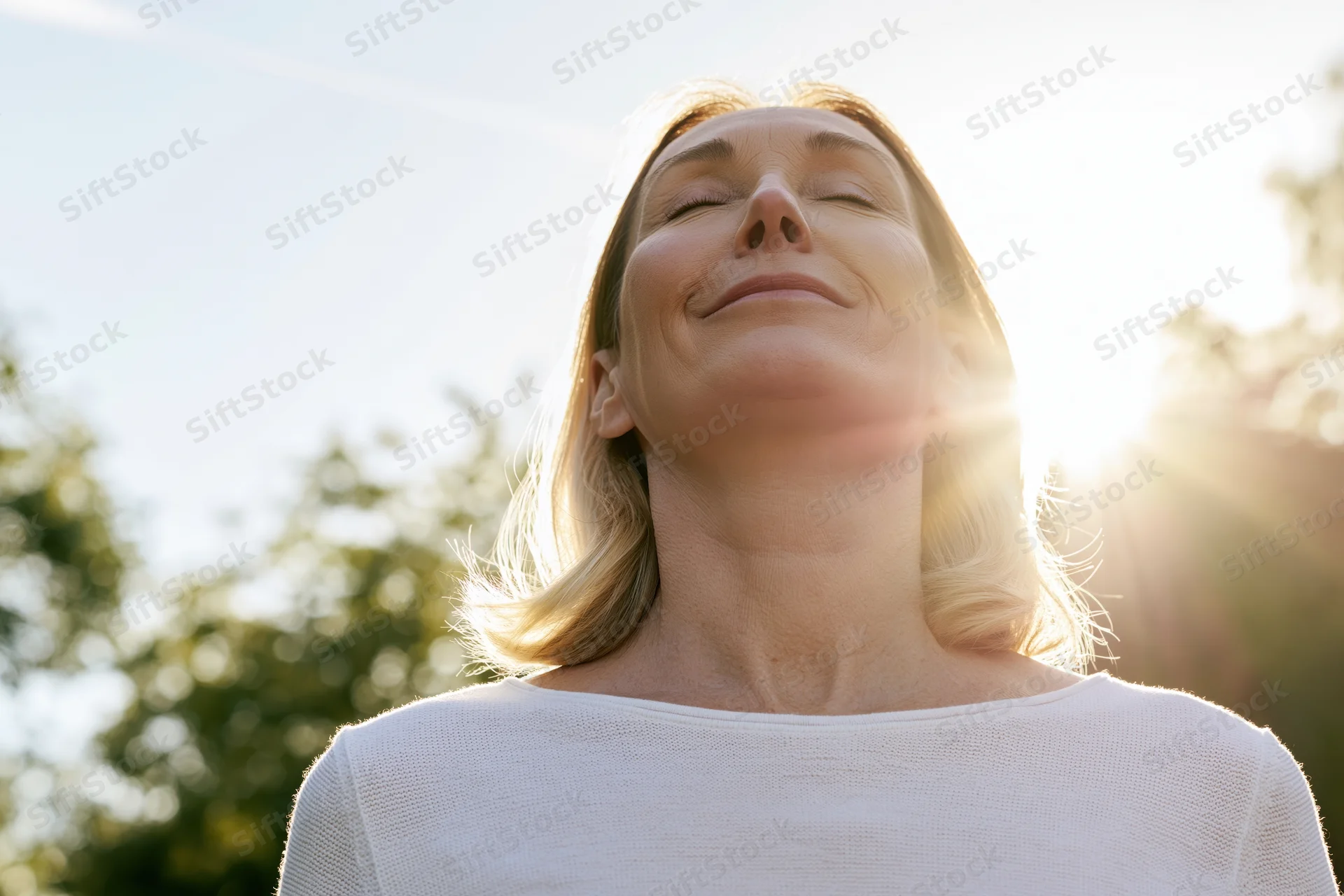 Middle-aged blonde woman tilting head back with eyes closed in sunlight outdoors