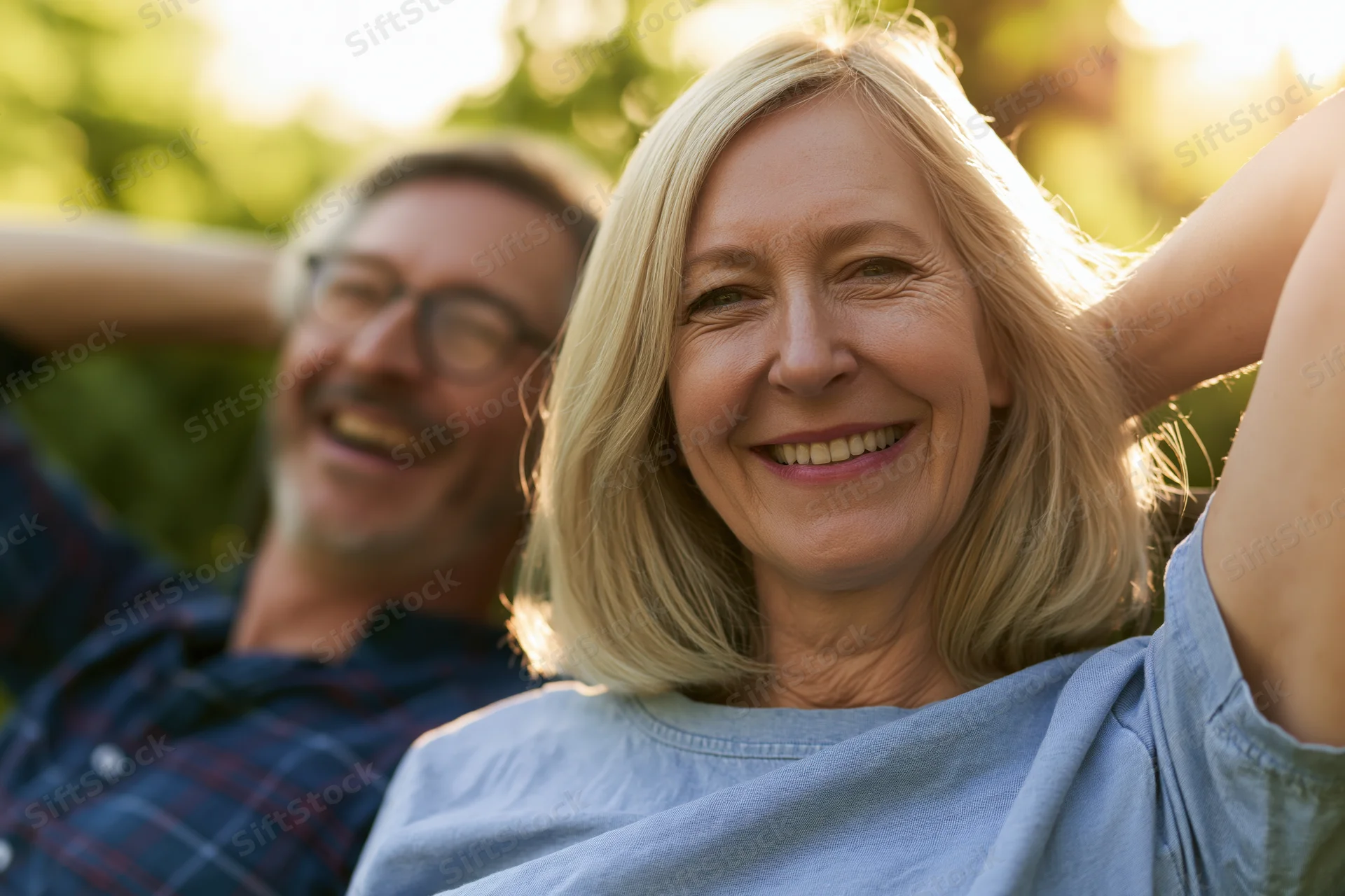 Middle-aged woman smiling with hands behind head, man in plaid shirt laughing behind her in a sunlit garden