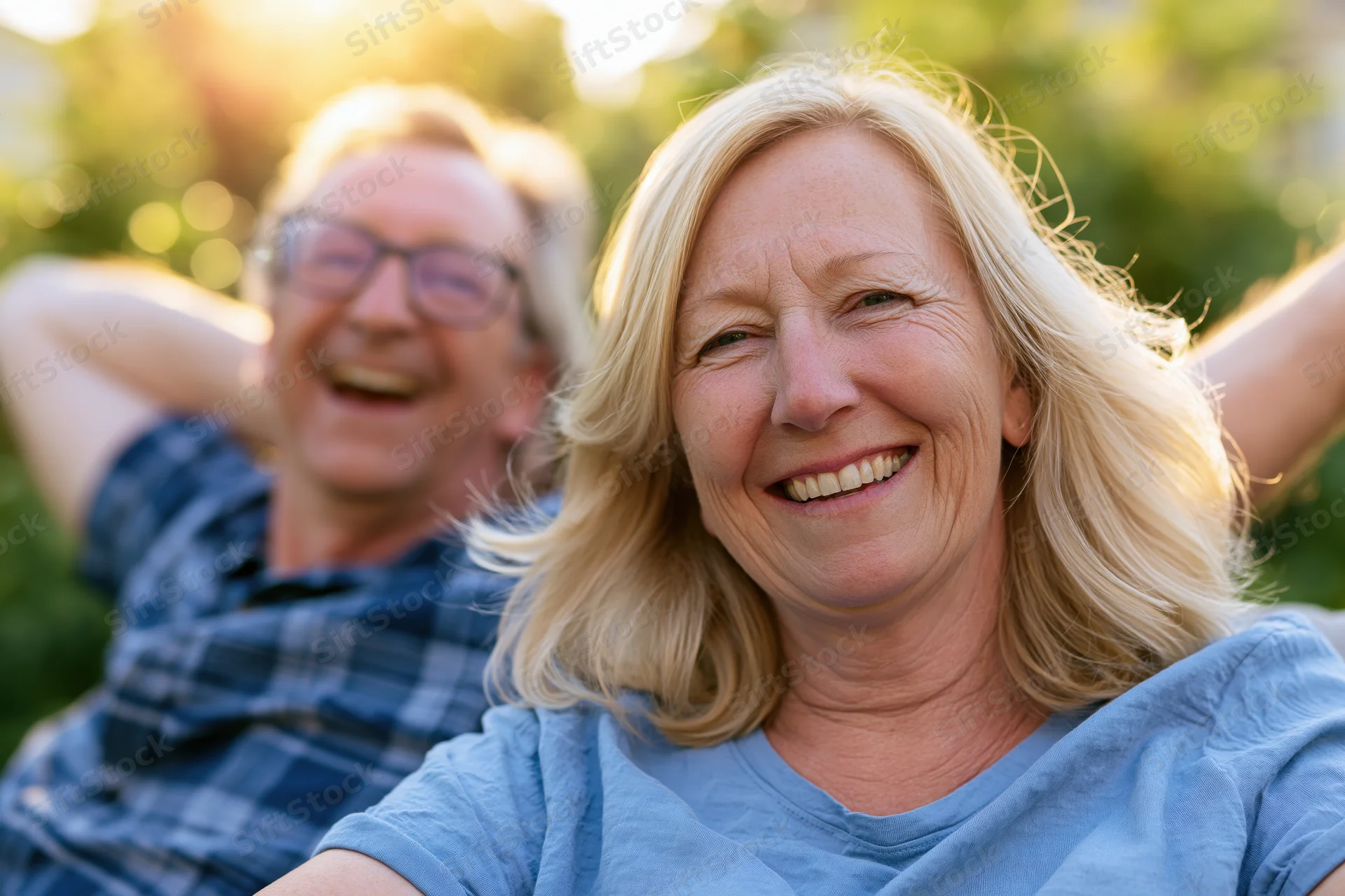 Smiling middle-aged woman with blonde hair sitting outdoors with a laughing man in the background