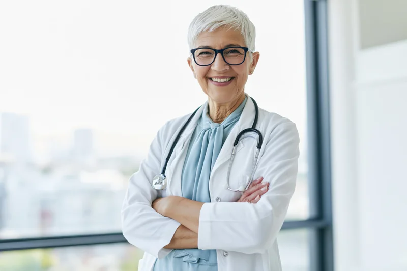 Senior female doctor in white coat with stethoscope smiling with arms crossed in clinic near window