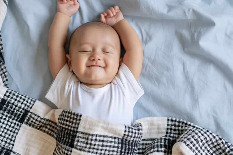 Baby sleeping with arms raised above head on bed covered with checkered blanket