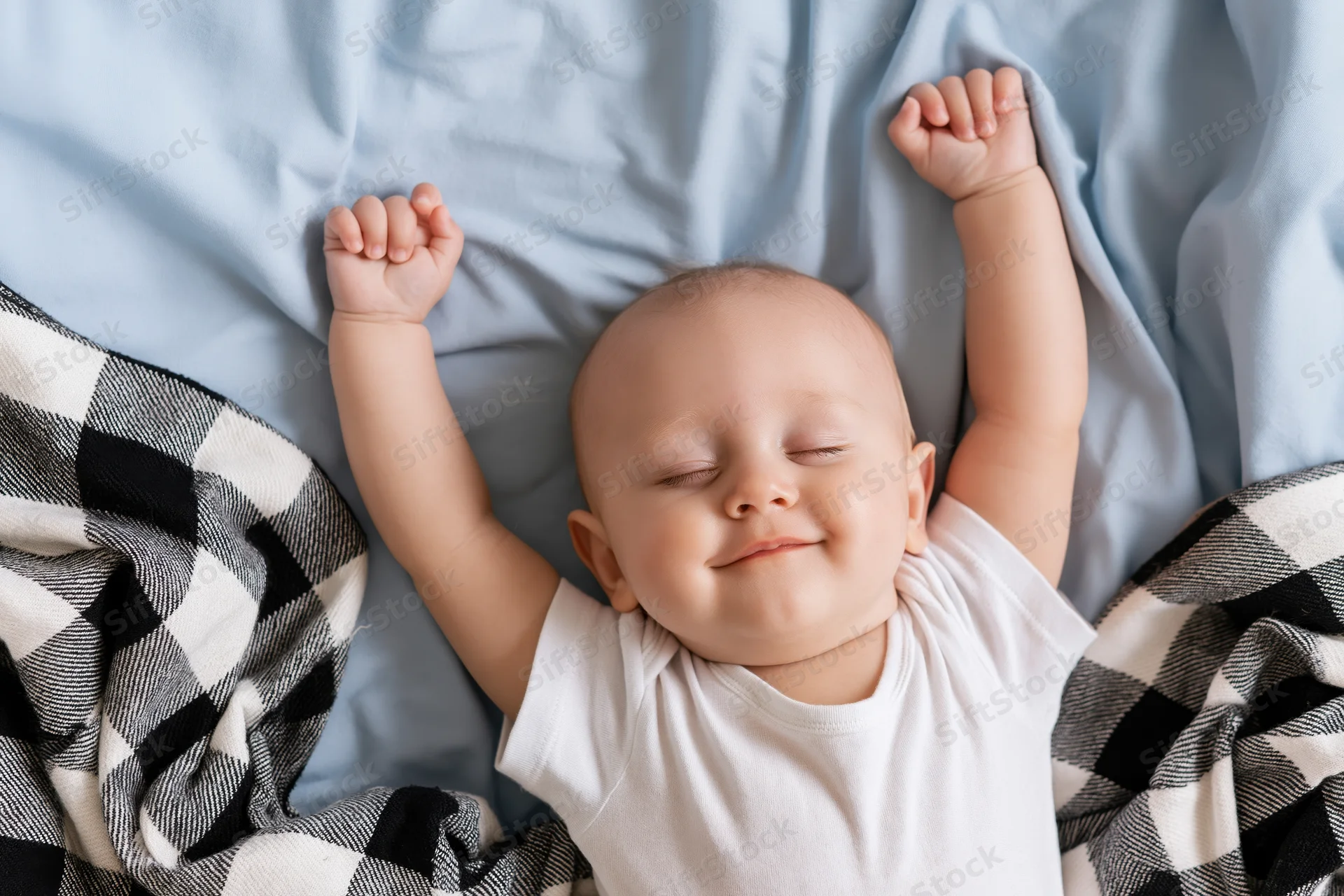 Baby sleeping with arms raised on a bed covered in a blue sheet and checkered blanket