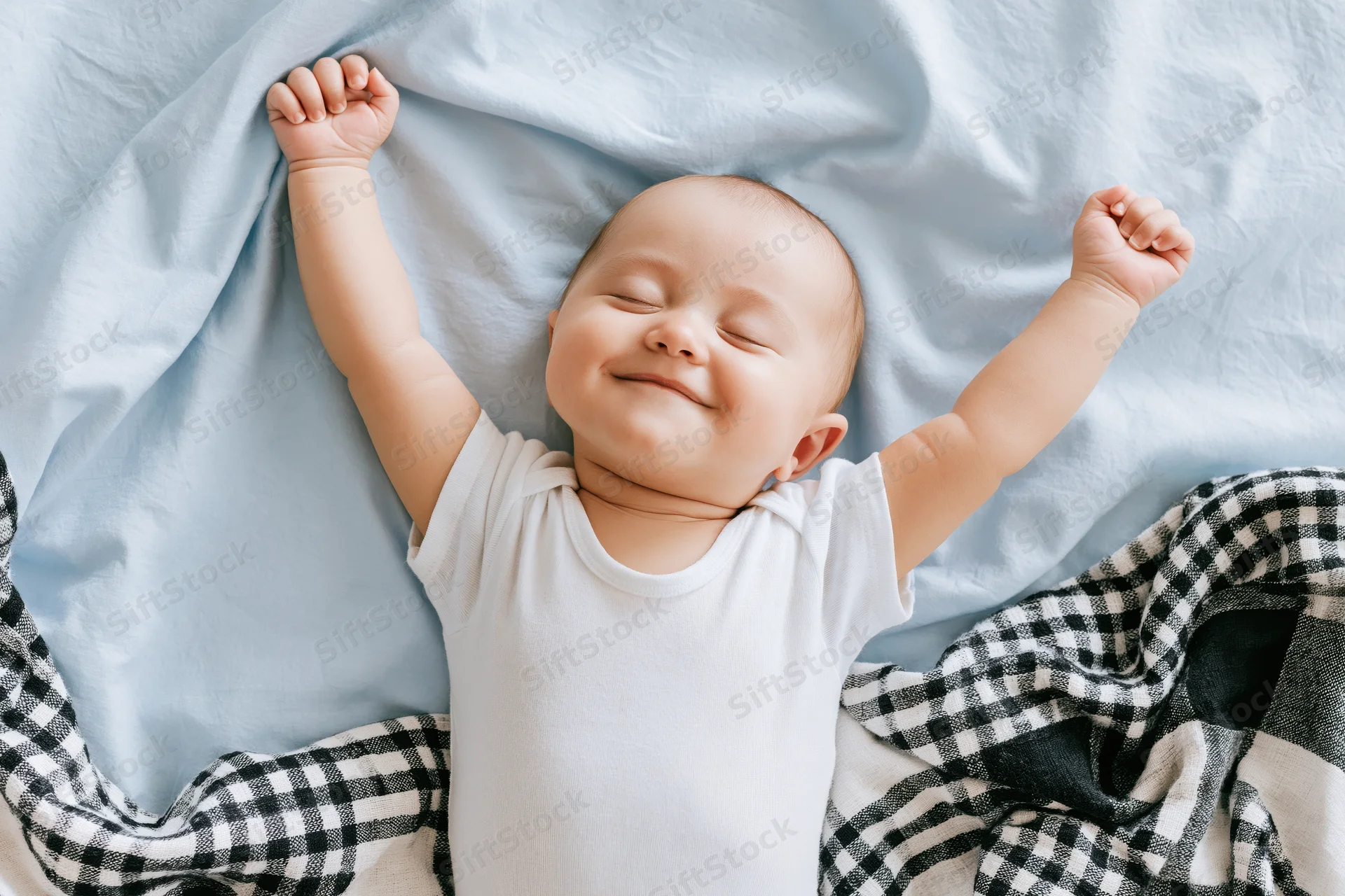 Smiling baby lying on a bed with arms raised above head, covered by a blue blanket and checkered fabric