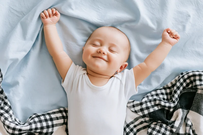 Smiling baby lying on a bed with arms raised above head, covered by a blue blanket and checkered fabric