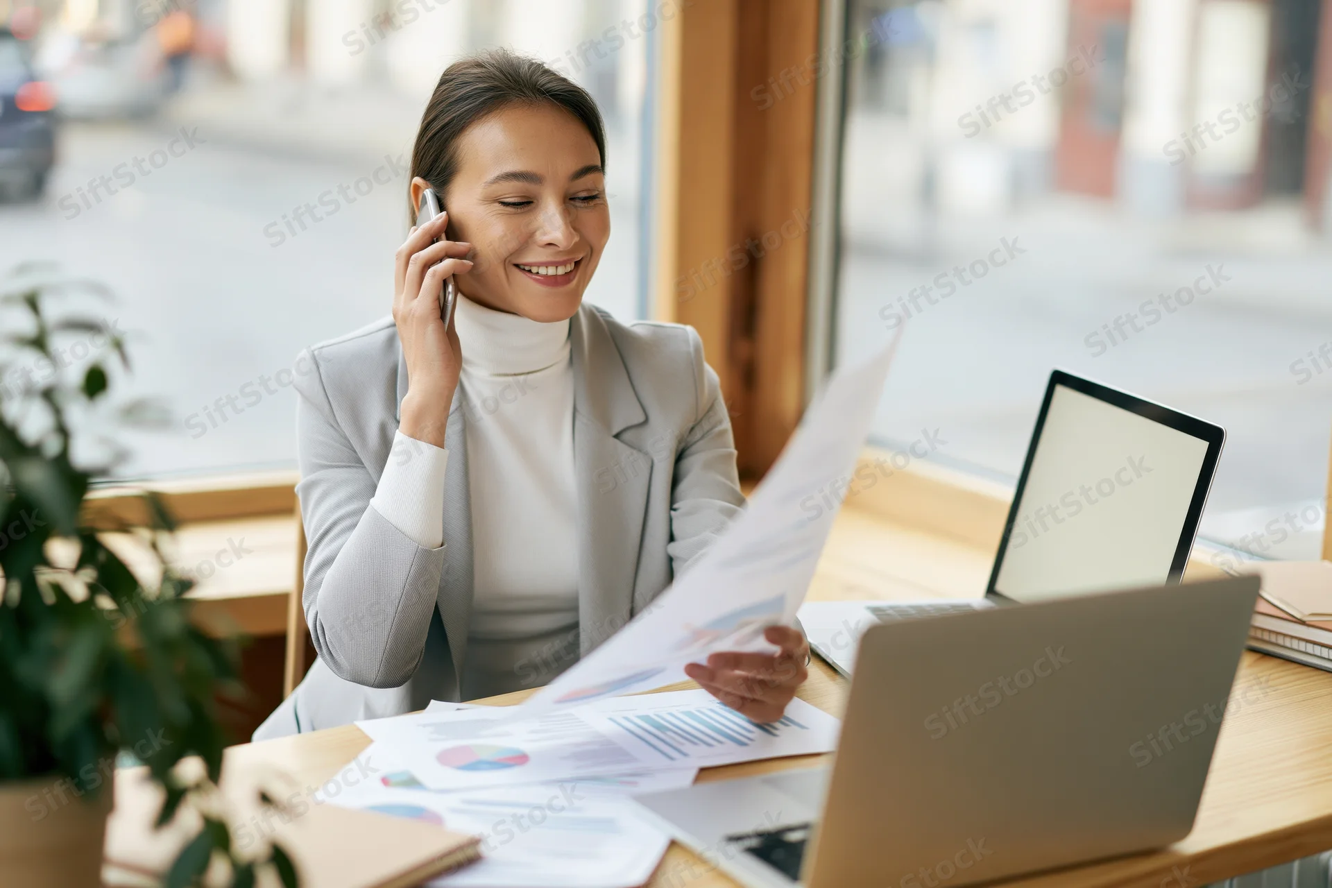 Smiling businesswoman talking on smartphone at office desk with documents and laptop