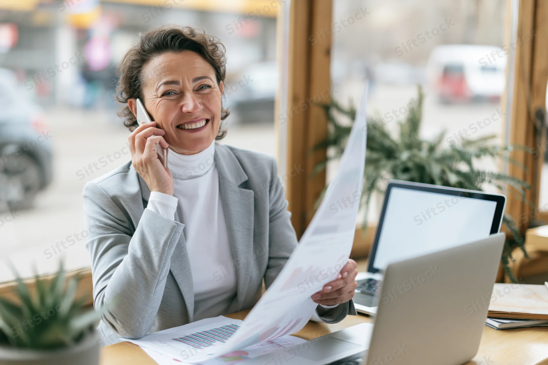 Smiling businesswoman talking on smartphone at office desk with documents and laptop