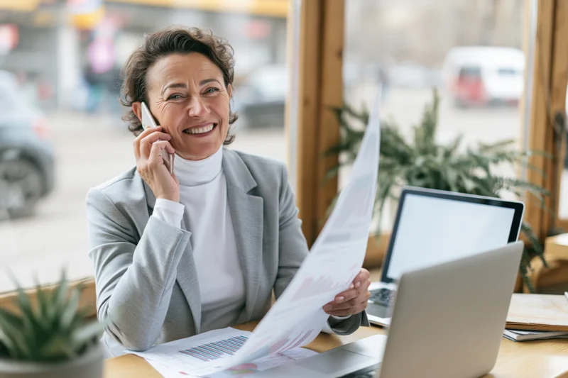 Smiling businesswoman talking on smartphone at office desk with documents and laptop