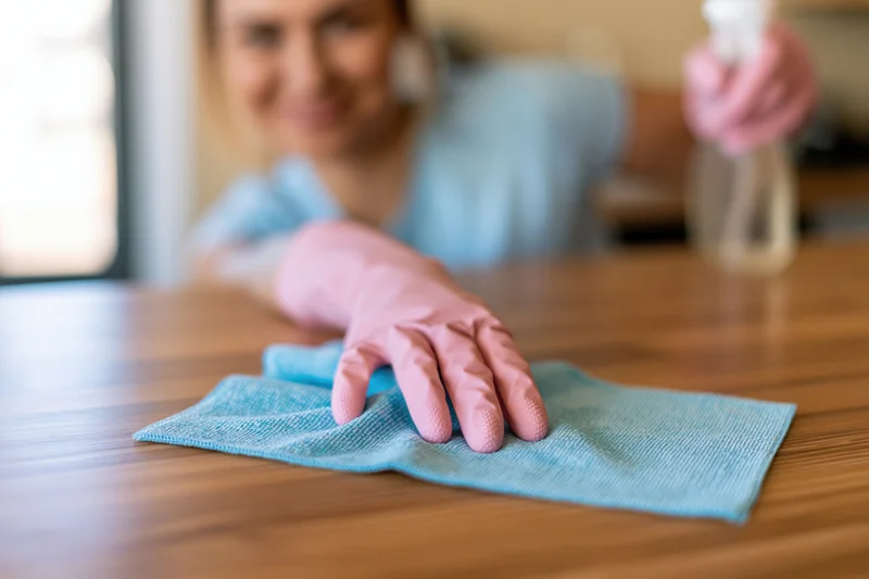 Woman wiping wooden table with blue microfiber cloth while holding spray bottle in background
