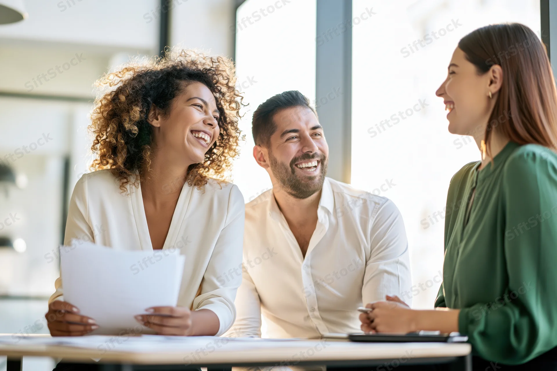 Three colleagues laughing during a casual office meeting at a desk with papers and a laptop