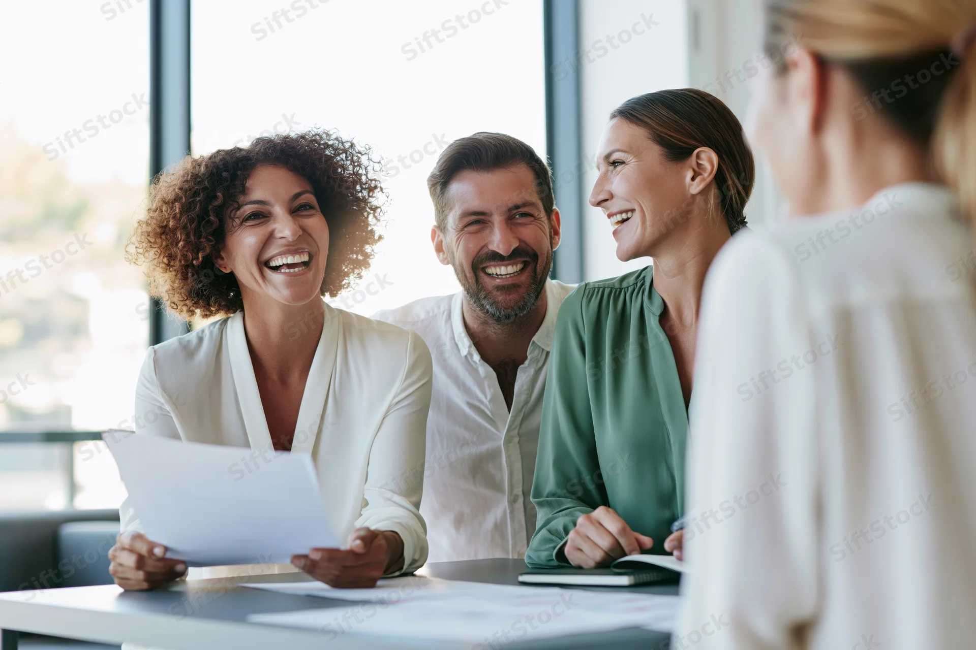 Colleagues laughing during a casual office meeting around a desk with documents