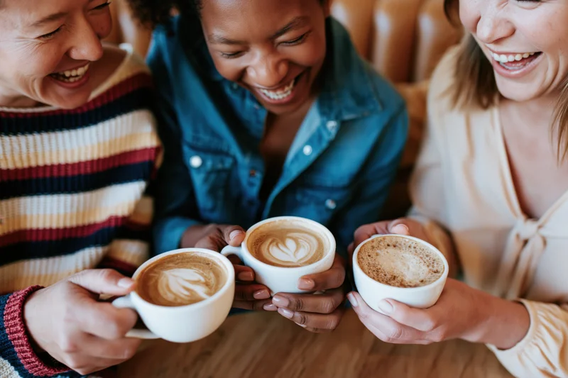 Three women laughing while holding cappuccinos at a café table
