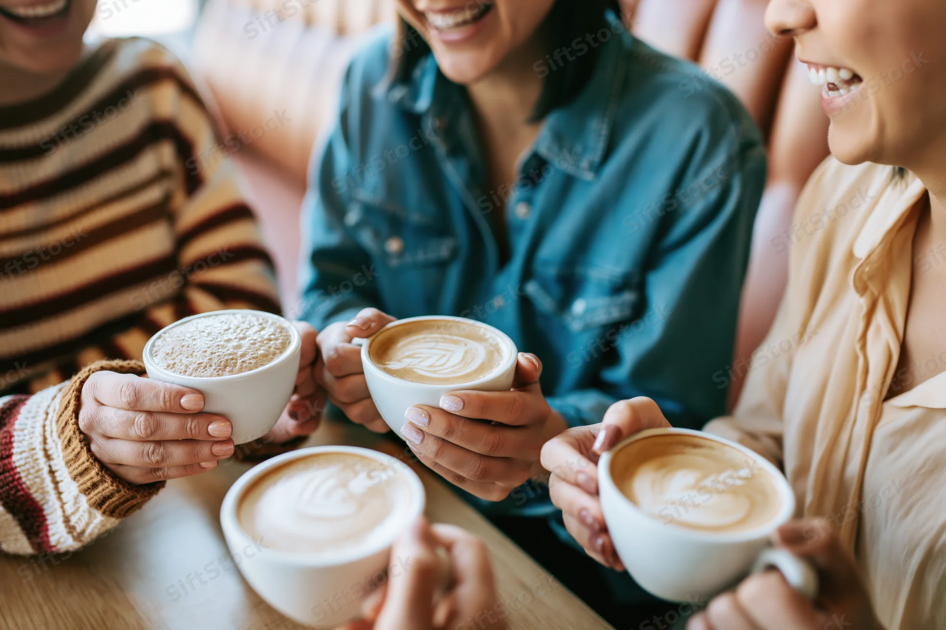 Three women laughing while holding cappuccinos at a café table