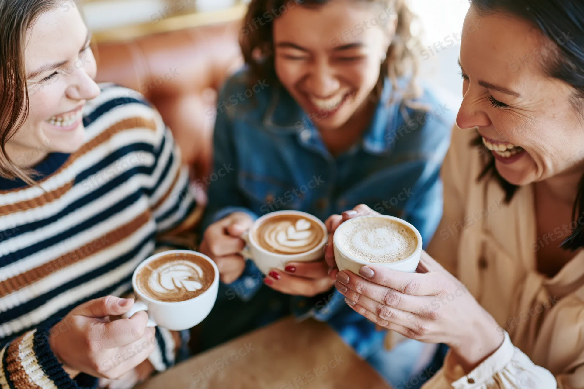 Three women laughing together at a café table holding cappuccinos