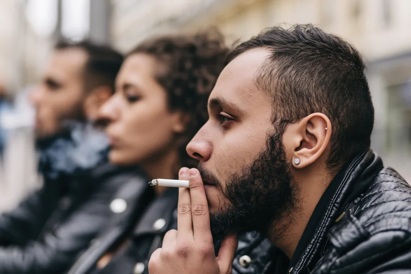 Man smoking a cigarette in profile with two friends in the background on a city sidewalk