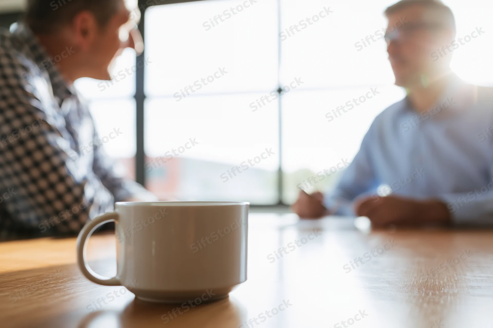 Two businessmen in conversation across a wooden meeting table with a coffee mug in focus