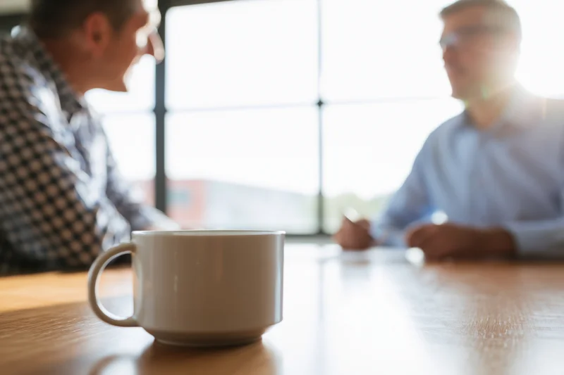 Two businessmen in conversation across a wooden meeting table with a coffee mug in focus